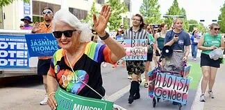 Older woman with white hair in a rainbow shirt and sunglasses waves at a Pride parade, holding a green campaign sign for Cyndi Munson for Governor.