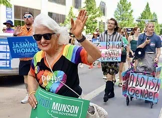 Oklahoma Pride Alliance looks beyond Pride Month Older woman with white hair in a rainbow shirt and sunglasses waves at a Pride parade, holding a green campaign sign for Cyndi Munson for Governor.