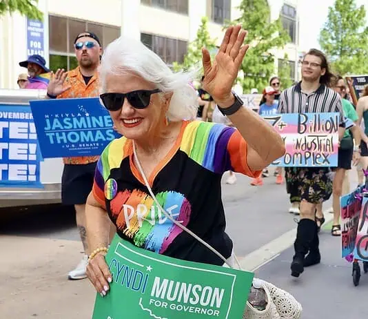 Oklahoma Pride Alliance looks beyond Pride Month Older woman with white hair in a rainbow shirt and sunglasses waves at a Pride parade, holding a green campaign sign for Cyndi Munson for Governor.