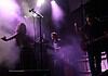 Live music performance on a foggy stage with purple lighting; female singer left, keyboardist center, guitarist right.
