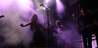 Live music performance on a foggy stage with purple lighting; female singer left, keyboardist center, guitarist right.