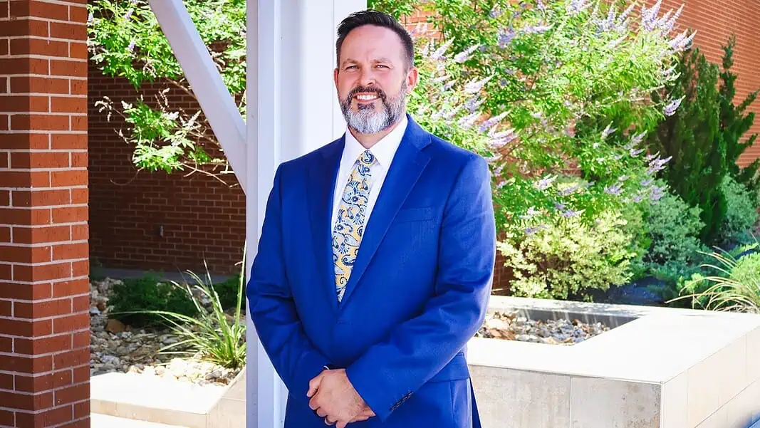 J. COWLEY METRO TECH Smiling man in a blue suit standing outdoors beside a brick column and greenery.