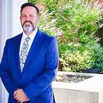 PRESS RELEASE: Metro Tech names Jeremy Cowley as new Deputy Superintendent Smiling man in a blue suit standing outdoors beside a brick column and greenery.