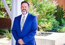 PRESS RELEASE: Metro Tech names Jeremy Cowley as new Deputy Superintendent Smiling man in a blue suit standing outdoors beside a brick column and greenery.