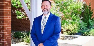 Smiling man in a blue suit standing outdoors beside a brick column and greenery.