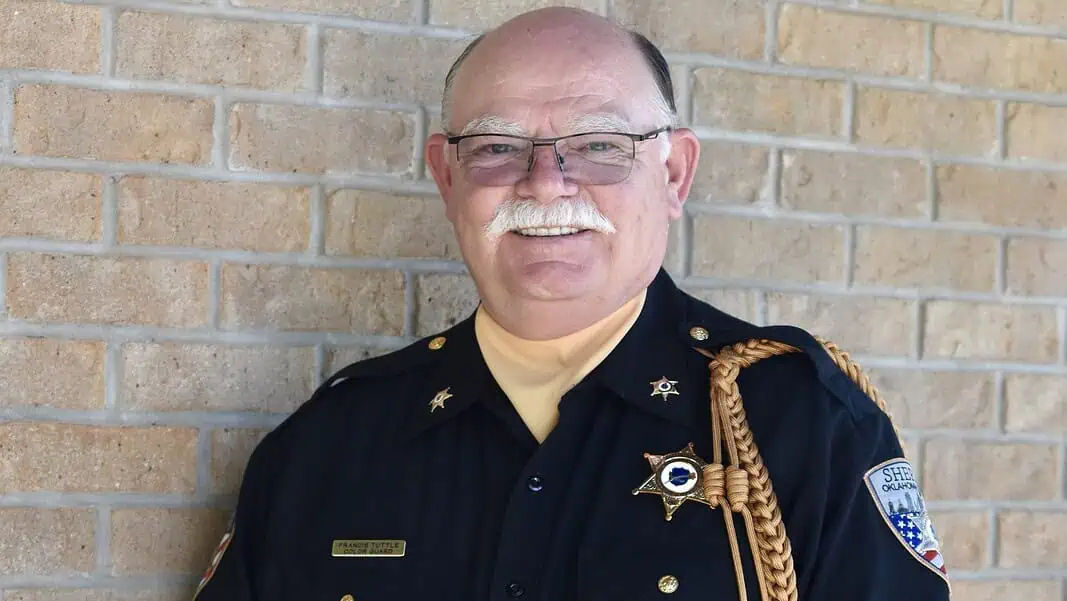 Johnnie Loudermilk Older male sheriff in uniform smiling against a brick wall, with gold braid and badge.