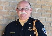 Older male sheriff in uniform smiling against a brick wall, with gold braid and badge.