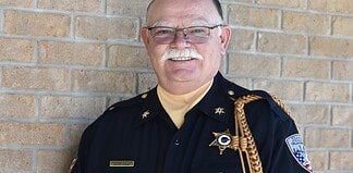 Older male sheriff in uniform smiling against a brick wall, with gold braid and badge.