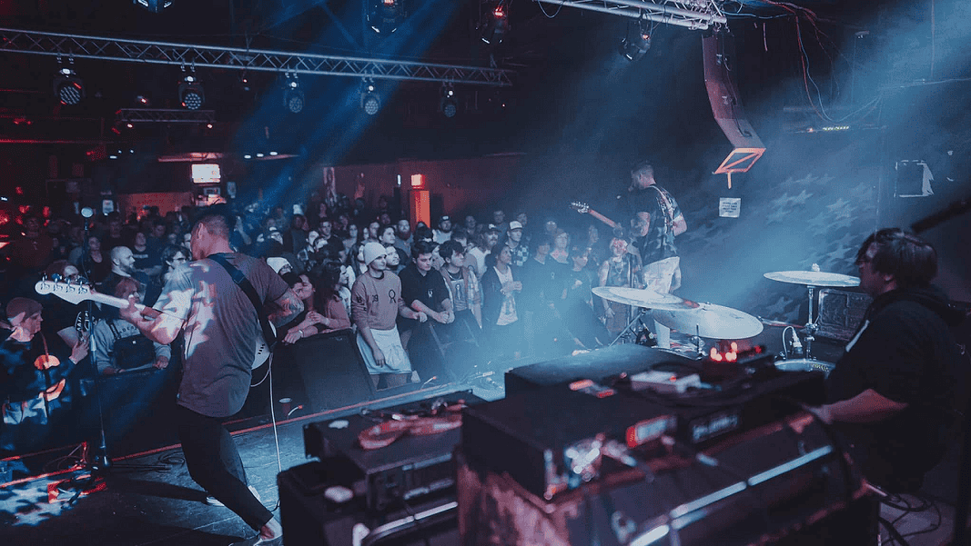 Lilac Kings Band performs on stage in a crowded club, guitarist at left, drummer on the right, blue and red stage lights illuminating the audience.