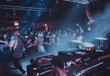 Showcases, mini-fests lift energy, noise, next generation of OKC music Band performs on stage in a crowded club, guitarist at left, drummer on the right, blue and red stage lights illuminating the audience.
