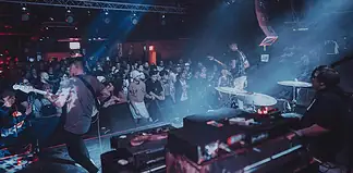 Band performs on stage in a crowded club, guitarist at left, drummer on the right, blue and red stage lights illuminating the audience.