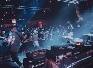 Showcases, mini-fests lift energy, noise, next generation of OKC music Band performs on stage in a crowded club, guitarist at left, drummer on the right, blue and red stage lights illuminating the audience.