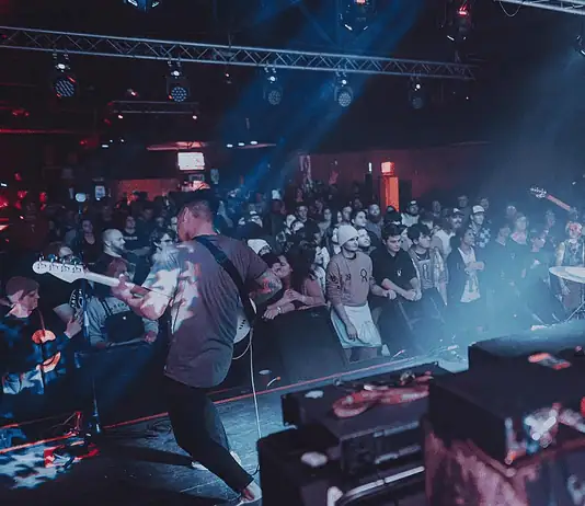 Showcases, mini-fests lift energy, noise, next generation of OKC music Band performs on stage in a crowded club, guitarist at left, drummer on the right, blue and red stage lights illuminating the audience.