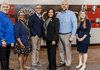 Six diverse professionals pose for a group photo in an office lobby with a red panel wall behind them.