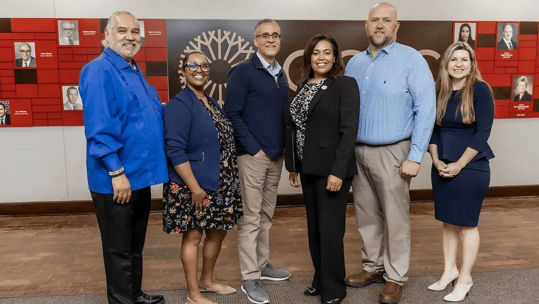 OCCC CoE award 2026 Six diverse professionals pose for a group photo in an office lobby with a red panel wall behind them.