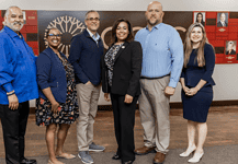 Six diverse professionals pose for a group photo in an office lobby with a red panel wall behind them.