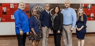Six diverse professionals pose for a group photo in an office lobby with a red panel wall behind them.