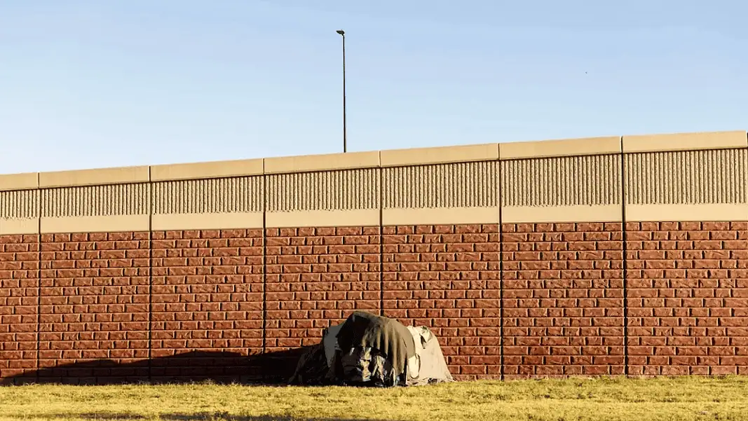 OK Watch homeless encampment A small tent covered with a tarp tucked against a long brick wall on a sunny day, with a grassy foreground.