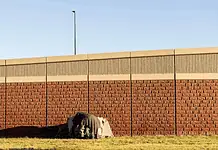 Three Cities, Three Approaches: How Oklahoma’s Homeless Encampment Sweeps Played Out Differently A small tent covered with a tarp tucked against a long brick wall on a sunny day, with a grassy foreground.