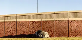 A small tent covered with a tarp tucked against a long brick wall on a sunny day, with a grassy foreground.