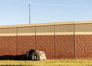 Three Cities, Three Approaches: How Oklahoma’s Homeless Encampment Sweeps Played Out Differently A small tent covered with a tarp tucked against a long brick wall on a sunny day, with a grassy foreground.