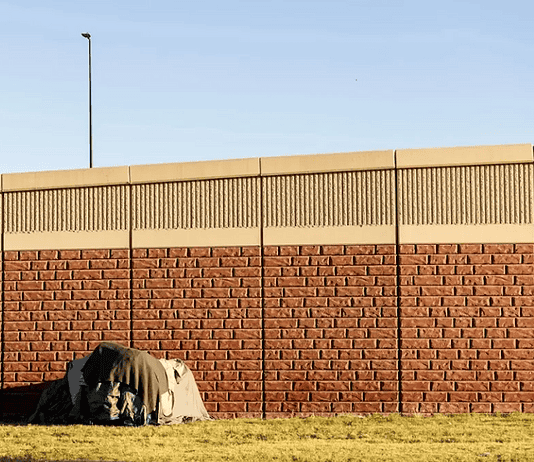 Three Cities, Three Approaches: How Oklahoma’s Homeless Encampment Sweeps Played Out Differently A small tent covered with a tarp tucked against a long brick wall on a sunny day, with a grassy foreground.