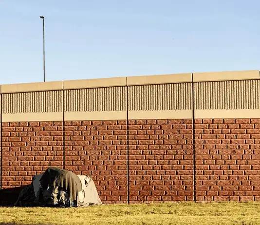Three Cities, Three Approaches: How Oklahoma’s Homeless Encampment Sweeps Played Out Differently A small tent covered with a tarp tucked against a long brick wall on a sunny day, with a grassy foreground.
