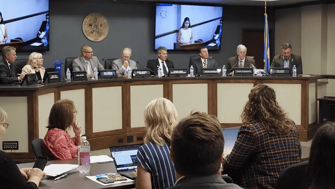 OTA OK Voice Panel of officials seated at a curved dais in a government hearing room, seal on the wall and large screens showing a speaker behind them, audience in foreground.