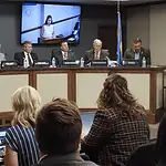 Oklahoma Turnpike Authority seeks $1.5B in bonds for construction costs Panel of officials seated at a curved dais in a government hearing room, seal on the wall and large screens showing a speaker behind them, audience in foreground.