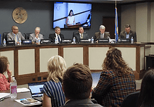 Panel of officials seated at a curved dais in a government hearing room, seal on the wall and large screens showing a speaker behind them, audience in foreground.