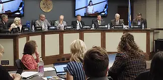 Panel of officials seated at a curved dais in a government hearing room, seal on the wall and large screens showing a speaker behind them, audience in foreground.