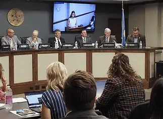 Oklahoma Turnpike Authority seeks $1.5B in bonds for construction costs Panel of officials seated at a curved dais in a government hearing room, seal on the wall and large screens showing a speaker behind them, audience in foreground.