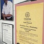 Wall exhibit with four large portraits of diverse adults, plus a yellow CHARTER poster labeled 'Youth Council' on a pink mat, and a framed council advisory list nearby, in a museum-like setting.