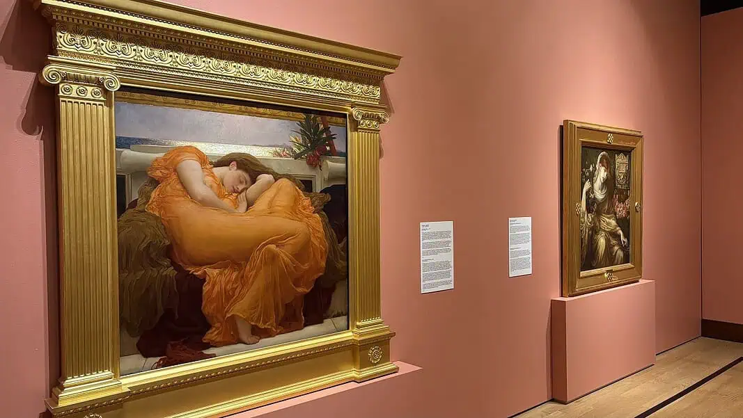 Photo Jun 18 2026, 9 54 49 AM Large ornate gold-framed painting on a pink wall, depicting a woman in an orange dress resting; to its right, a smaller framed religious-style artwork on a pedestal, also on pink wall.
