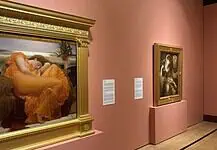 OKCMOA welcomes breadth, beauty of art from famed Puerto Rican museum Large ornate gold-framed painting on a pink wall, depicting a woman in an orange dress resting; to its right, a smaller framed religious-style artwork on a pedestal, also on pink wall.
