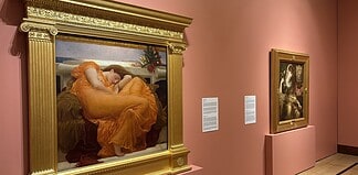 Large ornate gold-framed painting on a pink wall, depicting a woman in an orange dress resting; to its right, a smaller framed religious-style artwork on a pedestal, also on pink wall.