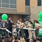 Group of people on a stage cutting a white ribbon at a grand opening, with green balloons behind them.