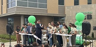 Group of people on a stage cutting a white ribbon at a grand opening, with green balloons behind them.