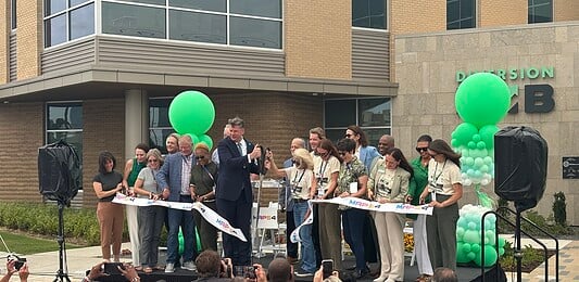 Group of people on a stage cutting a white ribbon at a grand opening, with green balloons behind them.