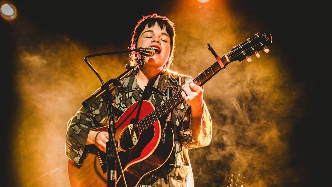Musician singing into a microphone while playing an acoustic guitar on stage with warm yellow lighting behind them.