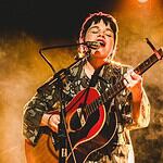 Simple, poignant power of songwriting on display in new releases Musician singing into a microphone while playing an acoustic guitar on stage with warm yellow lighting behind them.