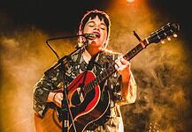 Musician singing into a microphone while playing an acoustic guitar on stage with warm yellow lighting behind them.