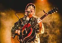 Simple, poignant power of songwriting on display in new releases Musician singing into a microphone while playing an acoustic guitar on stage with warm yellow lighting behind them.