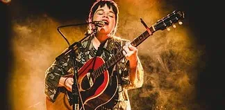 Musician singing into a microphone while playing an acoustic guitar on stage with warm yellow lighting behind them.