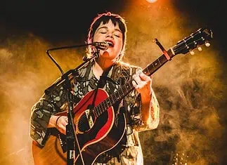 Simple, poignant power of songwriting on display in new releases Musician singing into a microphone while playing an acoustic guitar on stage with warm yellow lighting behind them.
