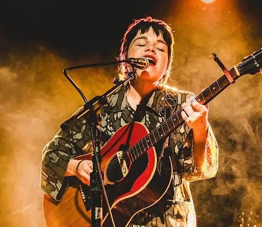 Simple, poignant power of songwriting on display in new releases Musician singing into a microphone while playing an acoustic guitar on stage with warm yellow lighting behind them.