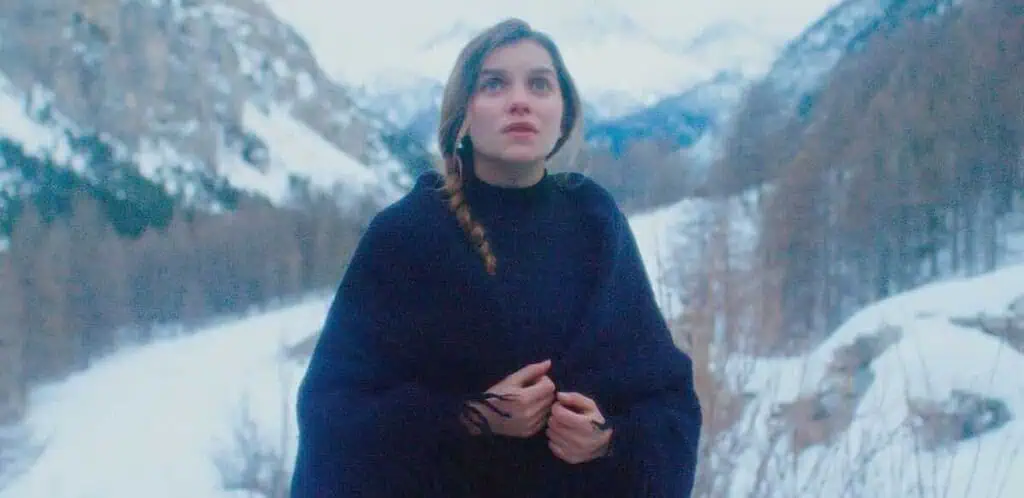 Woman in a dark poncho stands in a snowy mountain landscape, looking upward with a pensive expression.