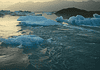Icebergs floating in a calm glacial lagoon at sunset with distant mountains in the background, reflecting on the water.