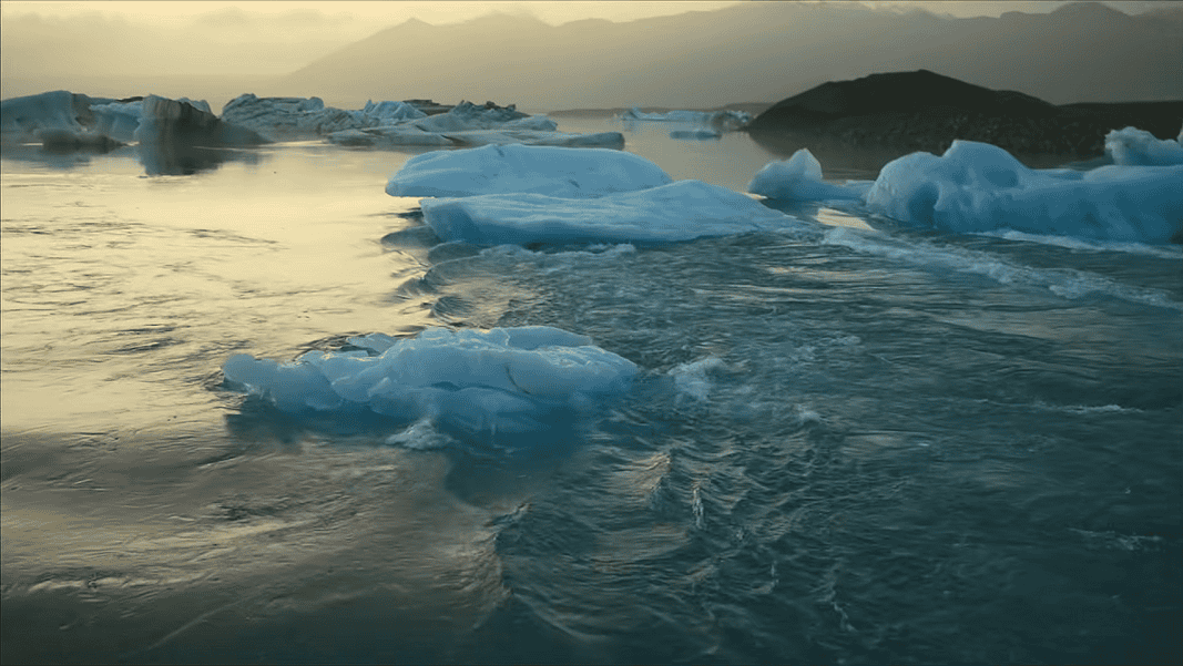 Icebergs floating in a calm glacial lagoon at sunset with distant mountains in the background, reflecting on the water.