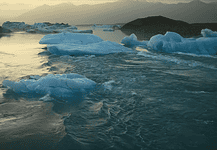 Icebergs floating in a calm glacial lagoon at sunset with distant mountains in the background, reflecting on the water.